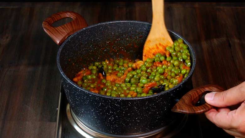 A wooden spatula is seen stirring the cooked green peas in a grey and white speckled non stick pan with two large wooden handles on an electric stove on a wooden countertop - Malvani Green Peas and Walnut Curry Recipe